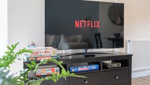 Ein Wohnzimmer mit einem großen Flachbildfernseher, der das Netflix-Logo anzeigt, einem Couchtisch mit Gesellschaftsspielen und einer dunkel gefärbten Unterhaltungsecke.