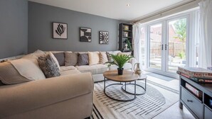 Ein modernes Wohnzimmer mit einem beigen Sofa, einem Couchtisch und einem Bücherregal. Drei gerahmte Kunstwerke sind an der Wand angebracht, und ein großes Fenster lässt Tageslicht herein.