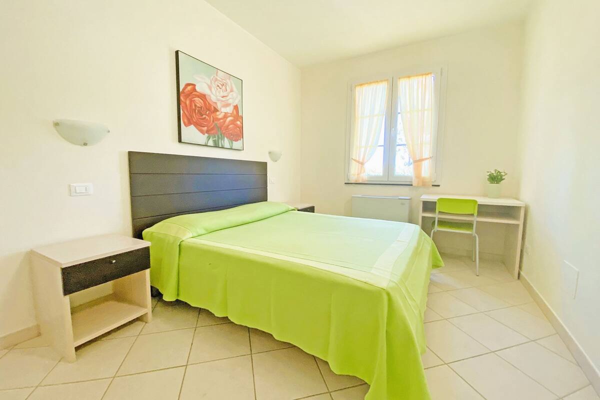 A bedroom with a bed, a desk, a chair, a nightstand, a wall-mounted lamp, and a window with curtains.