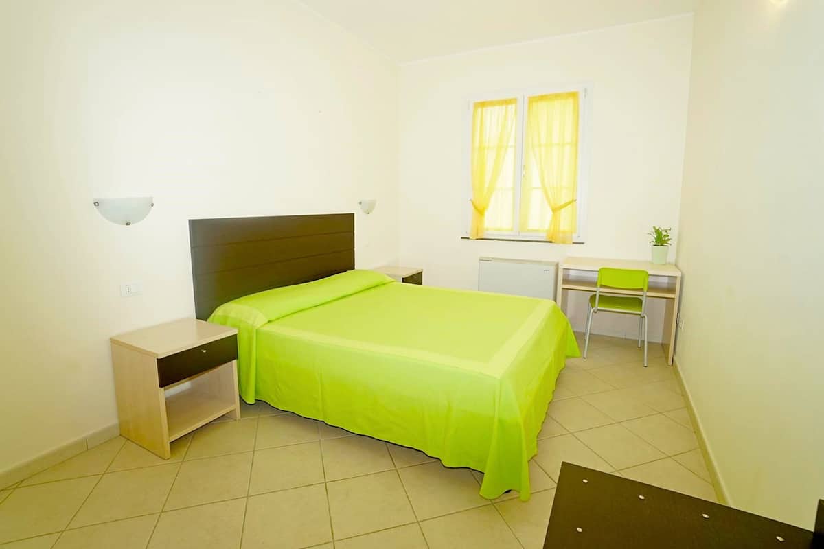 A small, bright room with a bed, a desk, and a chair.