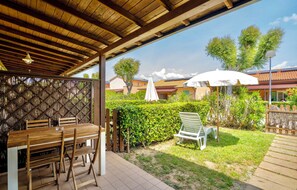 A covered outdoor seating area with a wooden table and chairs, a white umbrella, and a grassy lawn with a hedge. - Residence just 600 meters from the beach (Rosignano Marittimo)
