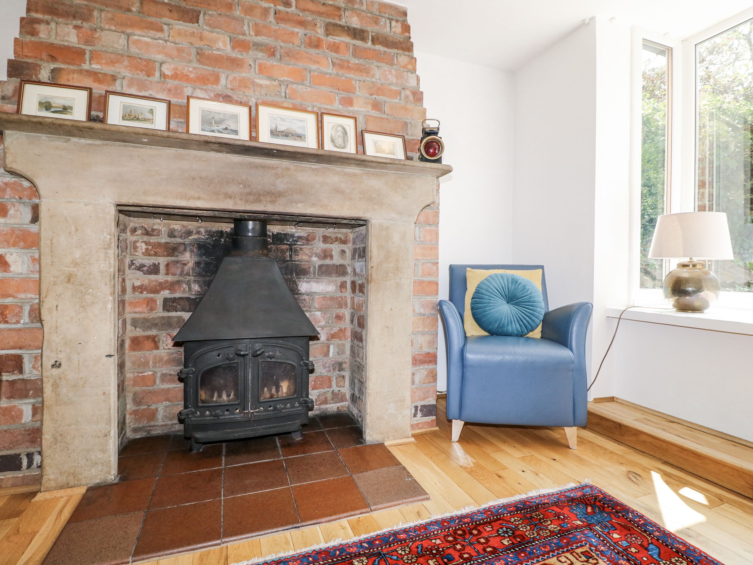 Una habitación con chimenea de ladrillo, un sillón azul, una alfombra estampada y una ventana con luz natural.