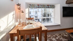 Un comedor con una mesa de madera dispuesta para cuatro personas, una ventana con un adornado de flores y un televisor montado en la pared.