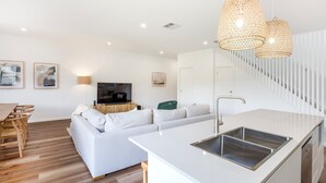 Una cocina moderna con una isla central, una zona de comedor con sillas de madera, un sofá blanco y una escalera que sube a un nivel superior.