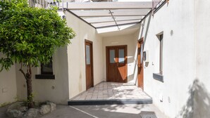 Un pasillo estrecho con piso de baldosas que conduce a una puerta de madera. Un pequeño árbol está plantado en una jardinera de rocas a la izquierda.