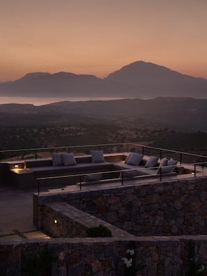 Una terraza en la azotea con un muro de piedra, un sofá con cojines y vistas a montañas y una masa de agua al atardecer.