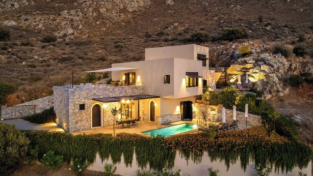 Uma casa moderna com piscina, paredes de pedra e vista para as colinas ao redor.