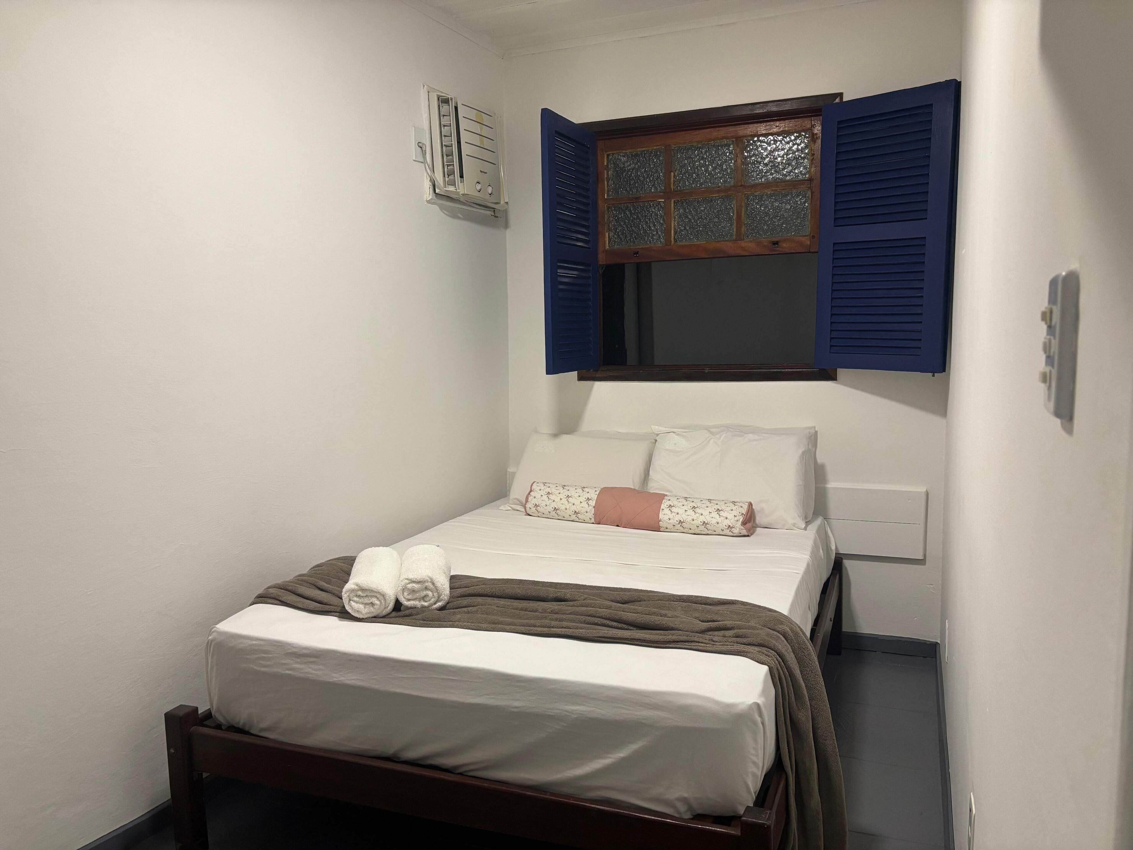 A small, neatly made bed with white linens and a brown blanket, a window with blue shutters, and a wall-mounted air conditioning unit.