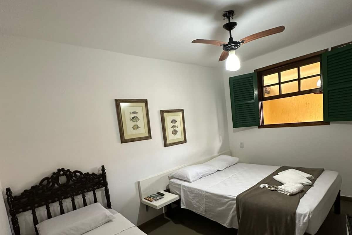 A room with two single beds, a ceiling fan, a window with shutters, and framed pictures on the wall.