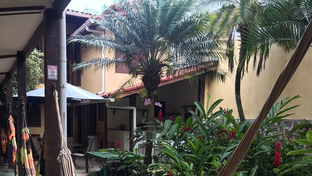 A hammock under a covered area with a stone-paved patio, surrounded by lush greenery and a building with a tiled roof. - Pousada Via Mar Buzios (Búzios)