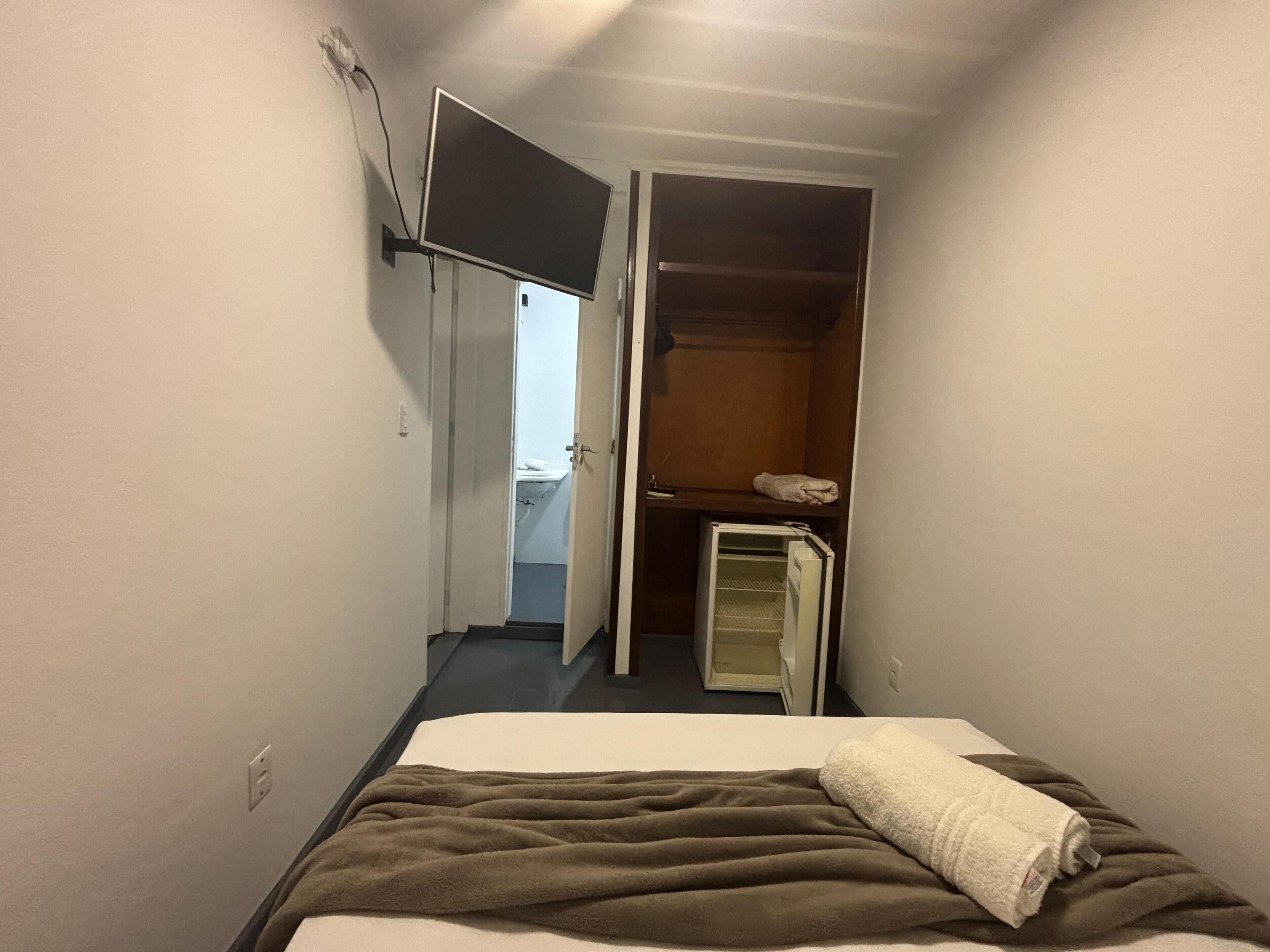 Una habitación de hotel estrecha con una cama, un armario y un refrigerador pequeño.
