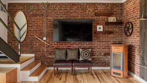 A room with a brick wall, a wooden bench with cushions, a television mounted above the bench, a small heater, and a staircase with wooden steps.