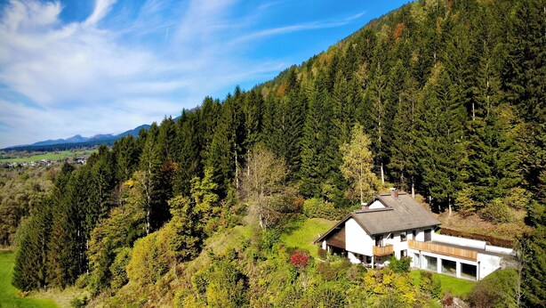 Una casa moderna con techo inclinado, rodeada de un bosque denso y montañas al fondo.