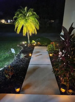 Uma passagem leva a um edifício, ladeada por plantas e iluminada por lanternas.