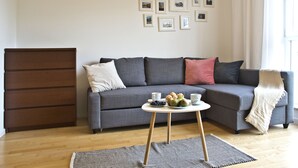 Ein Wohnzimmer mit grauem Sofa, einer Holzkommode, einem kleinen runden Tisch mit Essen und einer Wand mit gerahmten Bildern.
