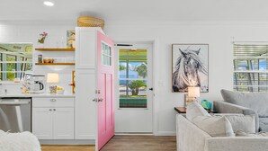A modern kitchen with white cabinets, a pink door, and a dining area with a grey sofa.