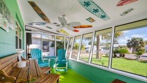A room with a wooden bench, a table, and two blue chairs. There is a ceiling fan and a view of the outside through large windows.
