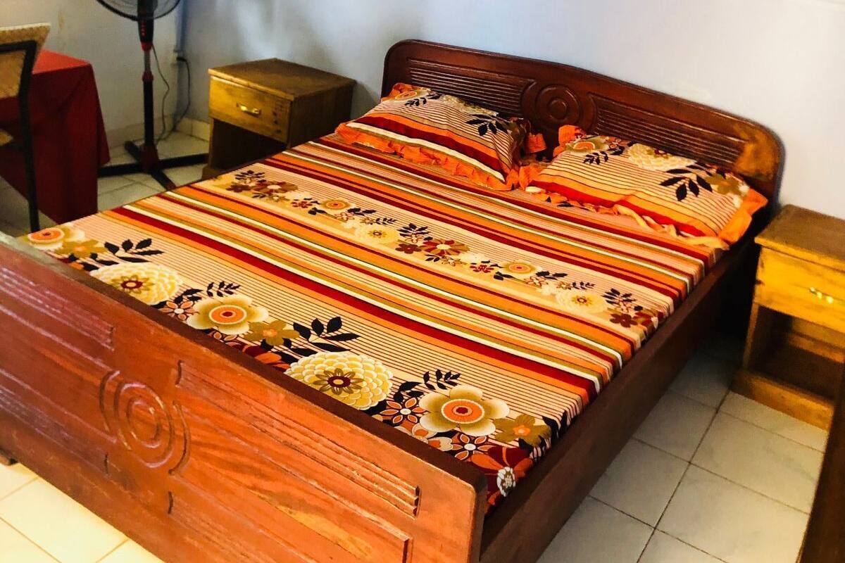 Uma cama de madeira com colcha florida, uma mesa de cabeceira, um criado-mudo, um ventilador fixado na parede e uma janela com cortinas.