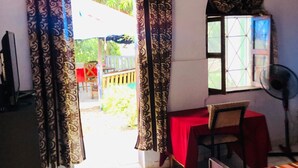 Una habitación con una cama, un escritorio con mantel rojo, una silla, una ventana con cortinas y un ventilador.