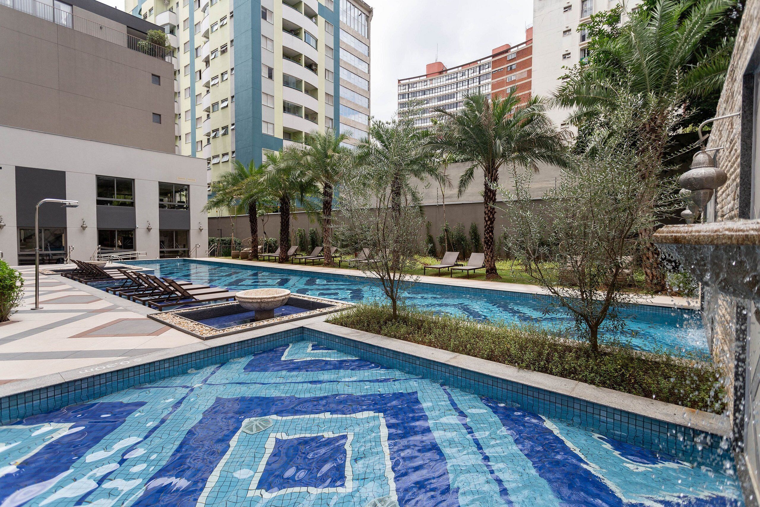 A swimming pool with mosaic tiles, surrounded by palm trees and benches, with high-rise buildings in the background.