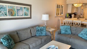 A living room with two grey sofas, a white coffee table with a decorative plate, a framed seashore art piece, a white lamp, and a kitchen area in the background. - Ground Floor Direct Oceanfront 2/2 (New Smyrna Beach)
