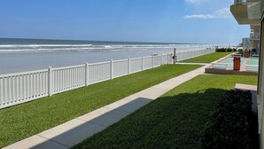 A beachfront property with a white fence, a grassy lawn, and a paved walkway leading to the beach. - Ground Floor Direct Oceanfront 2/2 (New Smyrna Beach)