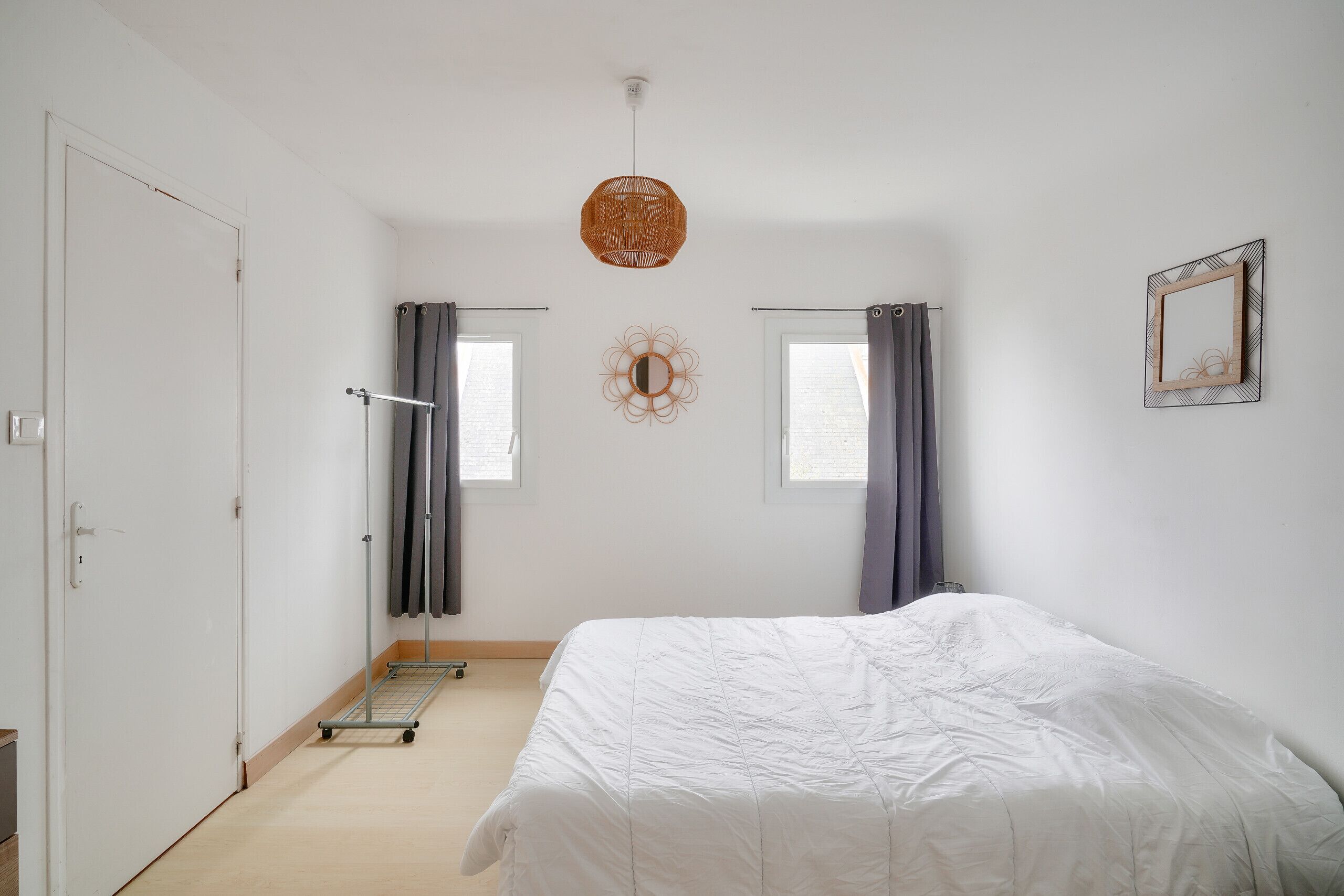 A bedroom with a bed, two windows with curtains, a wall-mounted mirror, and a ceiling light fixture.