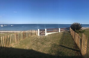 Une vue sur la côte, avec une clôture en bois menant à un portail, un ciel dégagé et des bateaux sur l’eau.