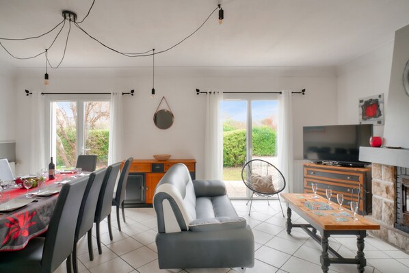 A modern living room with a sofa, dining table, and TV. There are large windows and a fireplace.