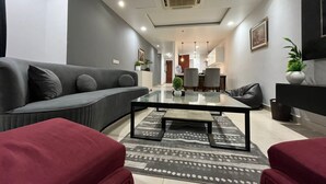 A modern living room with a grey sofa, a glass coffee table, and red armchairs. There is a dining area in the background with a kitchen visible.