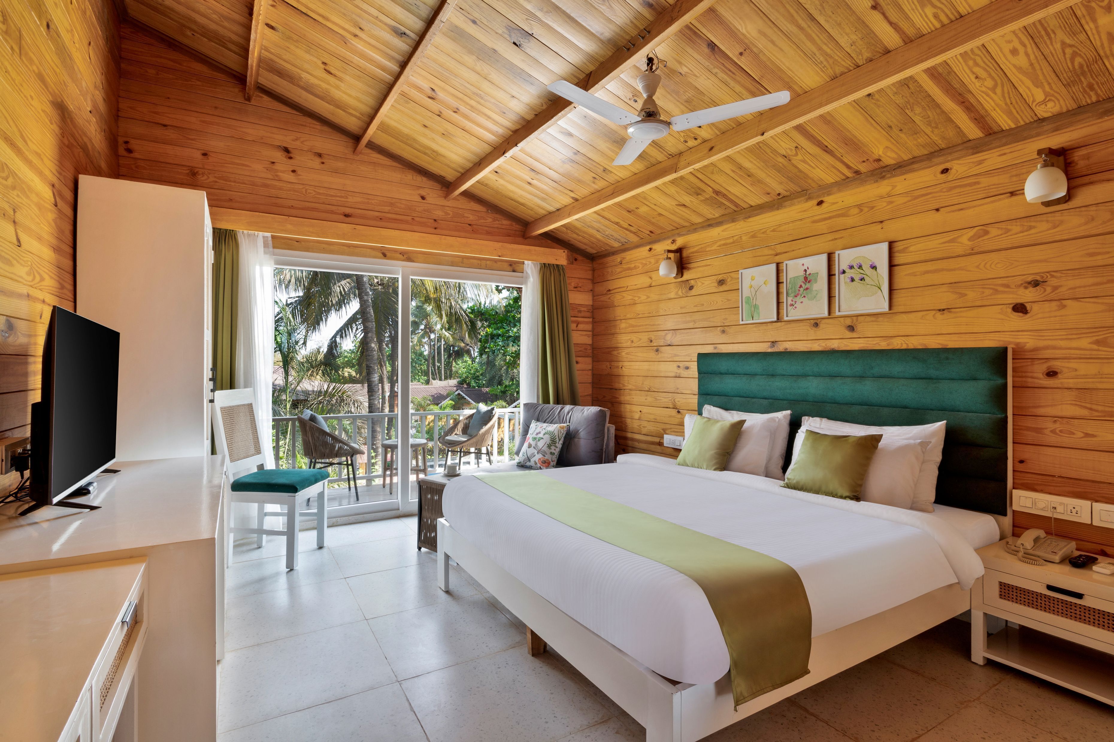 A wooden-paneled room with a bed, a desk, a chair, a TV, and a balcony with a view of palm trees.