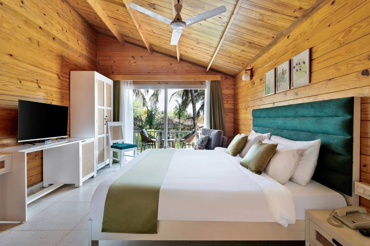 Une chambre de style cabane en bois, équipée d’un lit, d’une télévision et d’un balcon offrant une vue sur les palmiers.