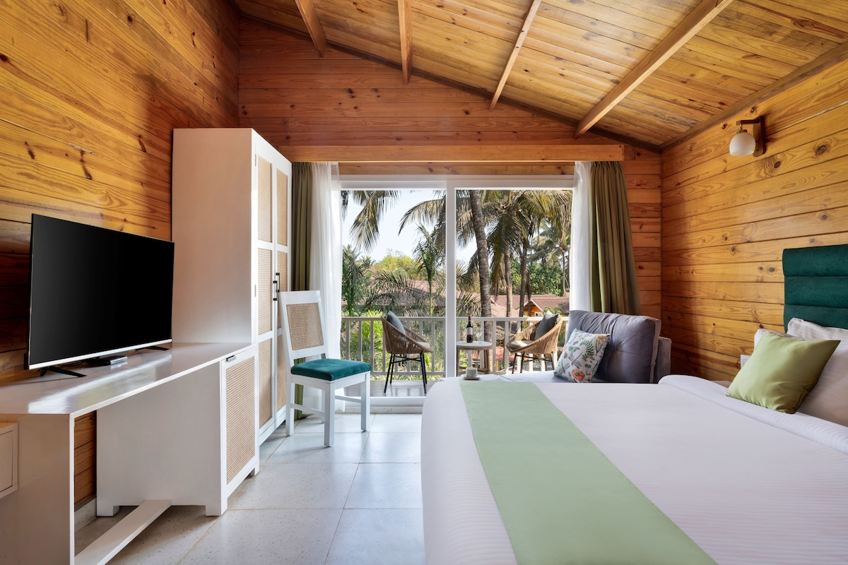 A wooden cabin room with a bed, a TV, a balcony, and a view of palm trees.
