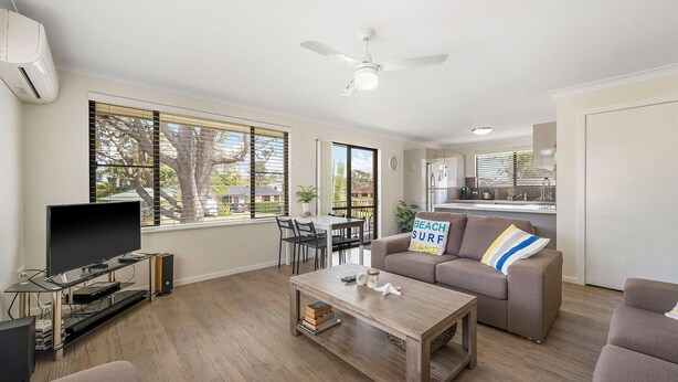A modern living room with a flat-screen TV, a wooden coffee table, a sofa with a beach-themed pillow, and a dining area with a table and chairs. - Bridgeview - Relaxing Getaway Near Surf Beach, Lake and Sand (Lake Cathie)