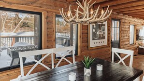 A wooden cabin interior with a dining table, white chairs, a chandelier, and large windows overlooking a snowy deck. - Beautiful Log Cabin, Pond adjacent on Big Bear Golf Course  (Big Bear Lake)
