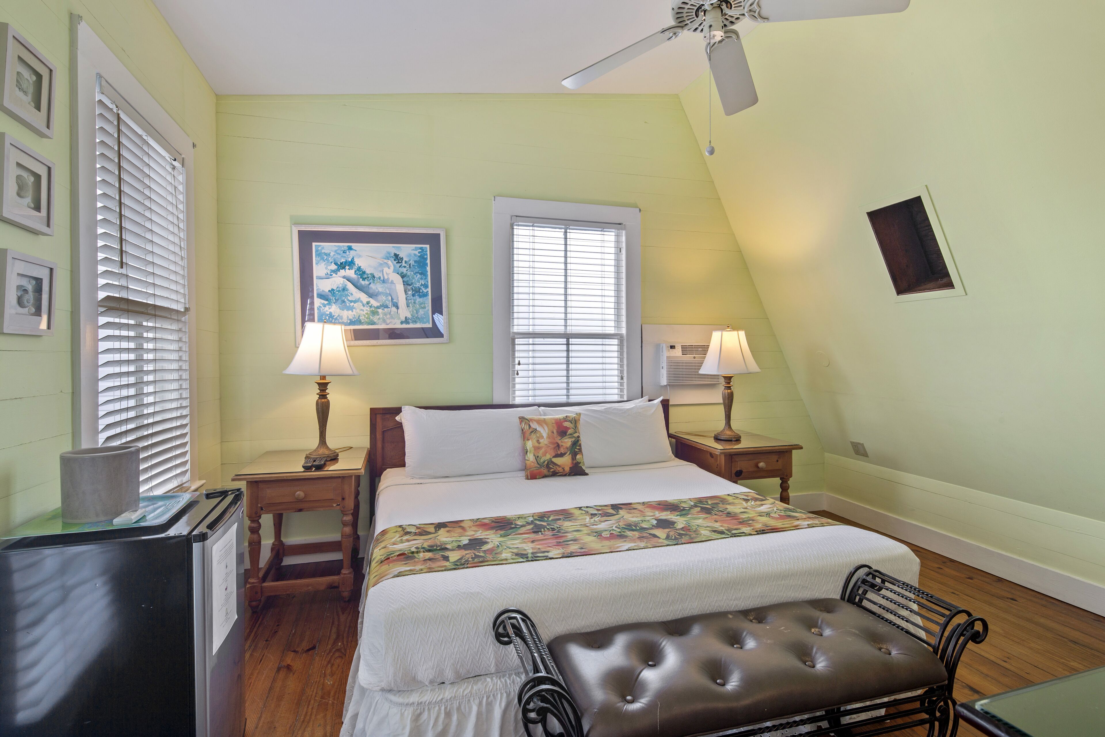 A bedroom with a bed, two bedside lamps, a ceiling fan, a window, and a small table.