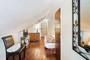 A bedroom with a wooden floor, a bed with a floral bedspread, a chair with a towel, a dresser, a desk with a lamp, and a mirror with a leaf design. - Curry House (Key West)
