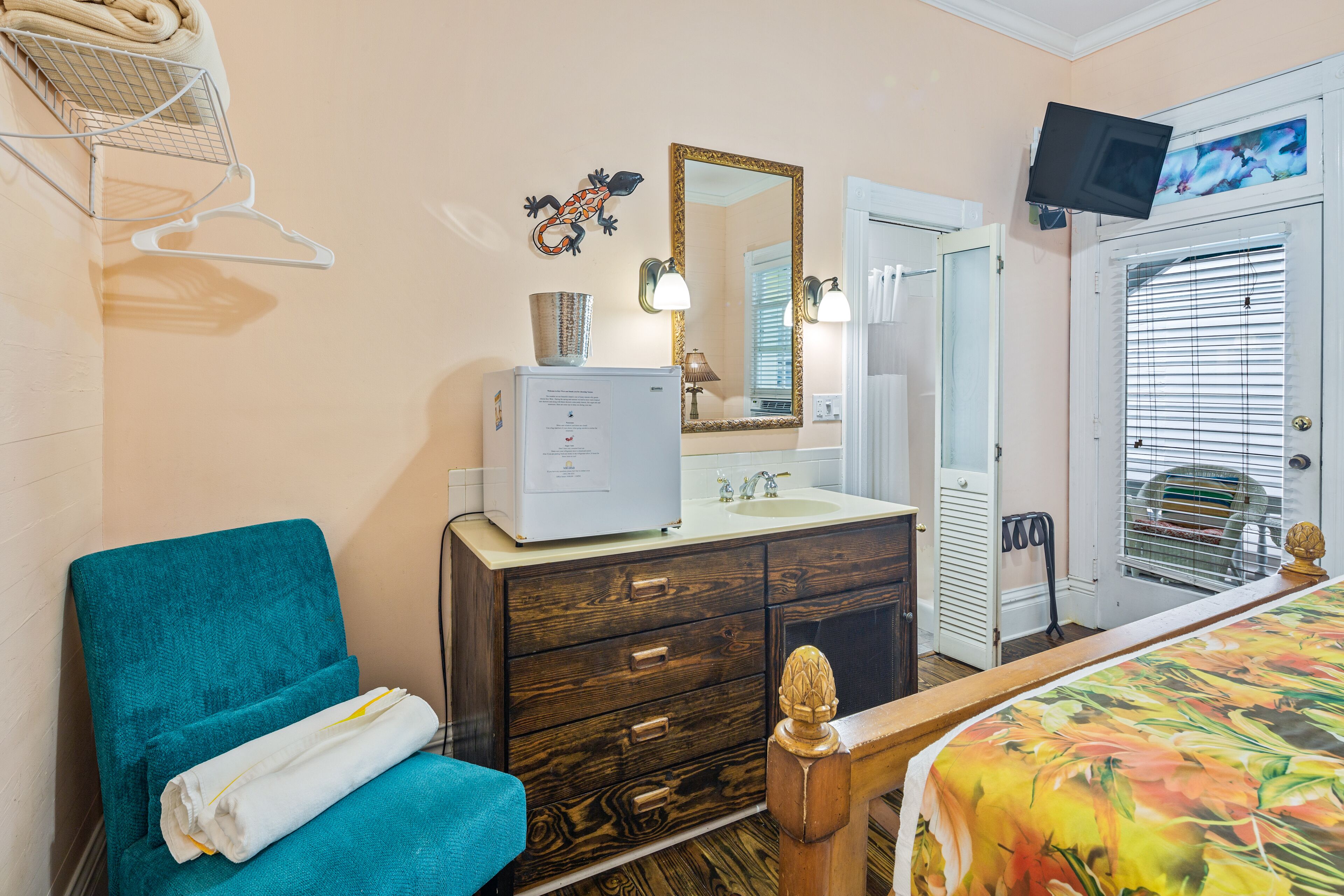 A bedroom with a teal chair, a wooden bed, a dresser with a mirror, and a television mounted on the wall.