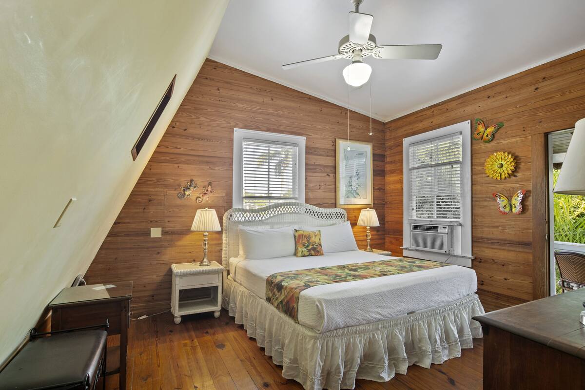 A bedroom with a sloped ceiling, wooden walls, a bed with a white headboard, two bedside tables with lamps, and a ceiling fan.