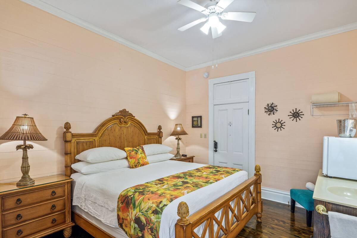 A bedroom with a wooden bed, two bedside lamps, a ceiling fan, a white door, and a small bathroom with a sink and mirror.