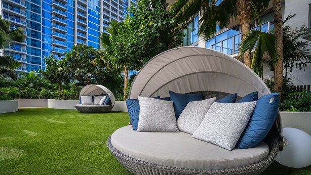 A circular outdoor seating area with cushions and a canopy, surrounded by lush greenery and a building in the background. - Newly Built, Hottest Condo at "Sky Ala Moana" 10th Floor  1 Bedroom (SAW1005) (Honolulu)