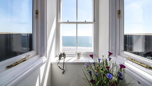 A room with a large window overlooking the ocean, a white wall, and a small window with a brass handle. - The Penthouse - Luxury 1 Bed - Panorama - Tenby (Tenby)