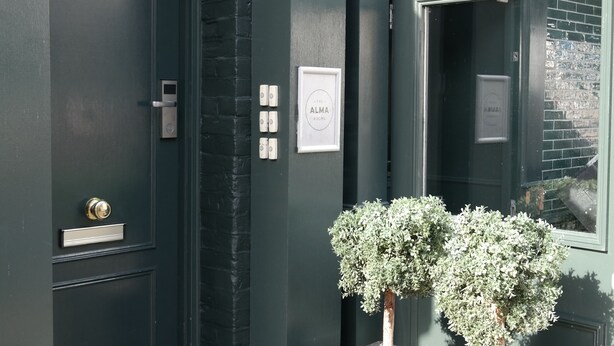 Un bâtiment de couleur vert foncé, doté d’une porte, d’une fenêtre et de deux plantes en pot.