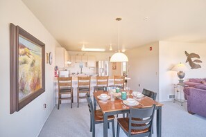A dining area with a wooden table set for four, surrounded by wooden chairs. A kitchen is visible in the background with white cabinets and a hanging light fixture. - Amazing Ocean View, In Town, Walk to Beach (Top) (Long Beach)