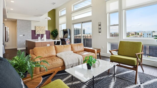 A spacious living area with a leather sofa, a green armchair, and a coffee table. There is a large window providing a view of the cityscape. - Skyline Penthouse | Espadin LoHi (Denver)