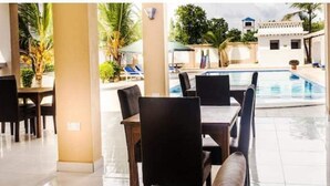 Zona junto a la piscina con mesa y sillas, una piscina y vista a un edificio de fachada blanca.