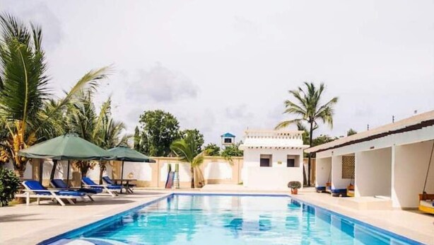 Una piscina con sillas de descanso y sombrillas, rodeada de palmeras y un edificio pequeño al fondo.