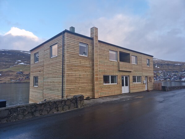 A two-story wooden building with a stone wall in front, near a body of water. - Guesthouse Hugo (Sorvagur)