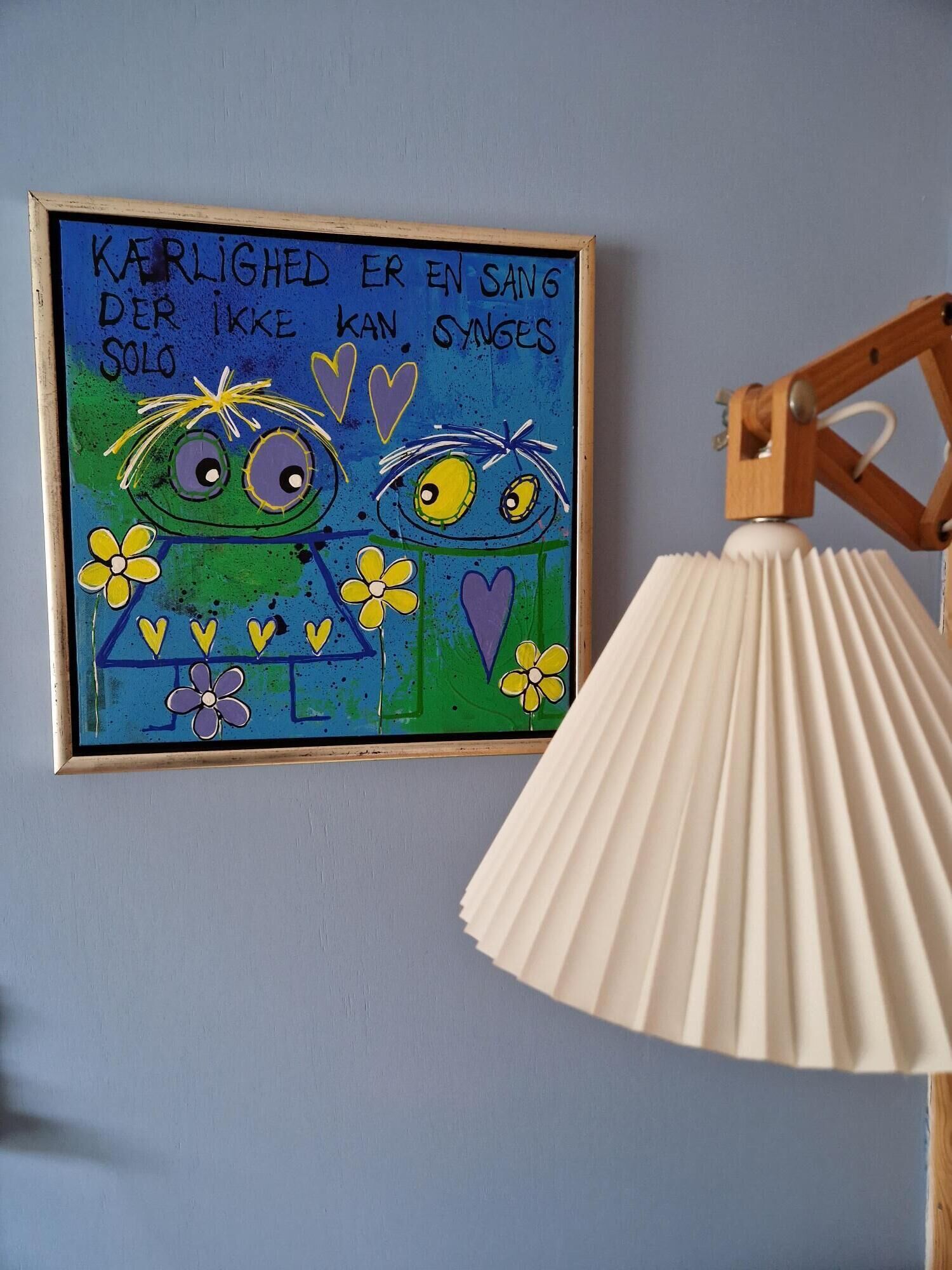 Et indrammet kunstværk med to tegneserieagtige ansigter og tekst på et fremmedsprog, en hvid plisseret lampe og en blå væg.