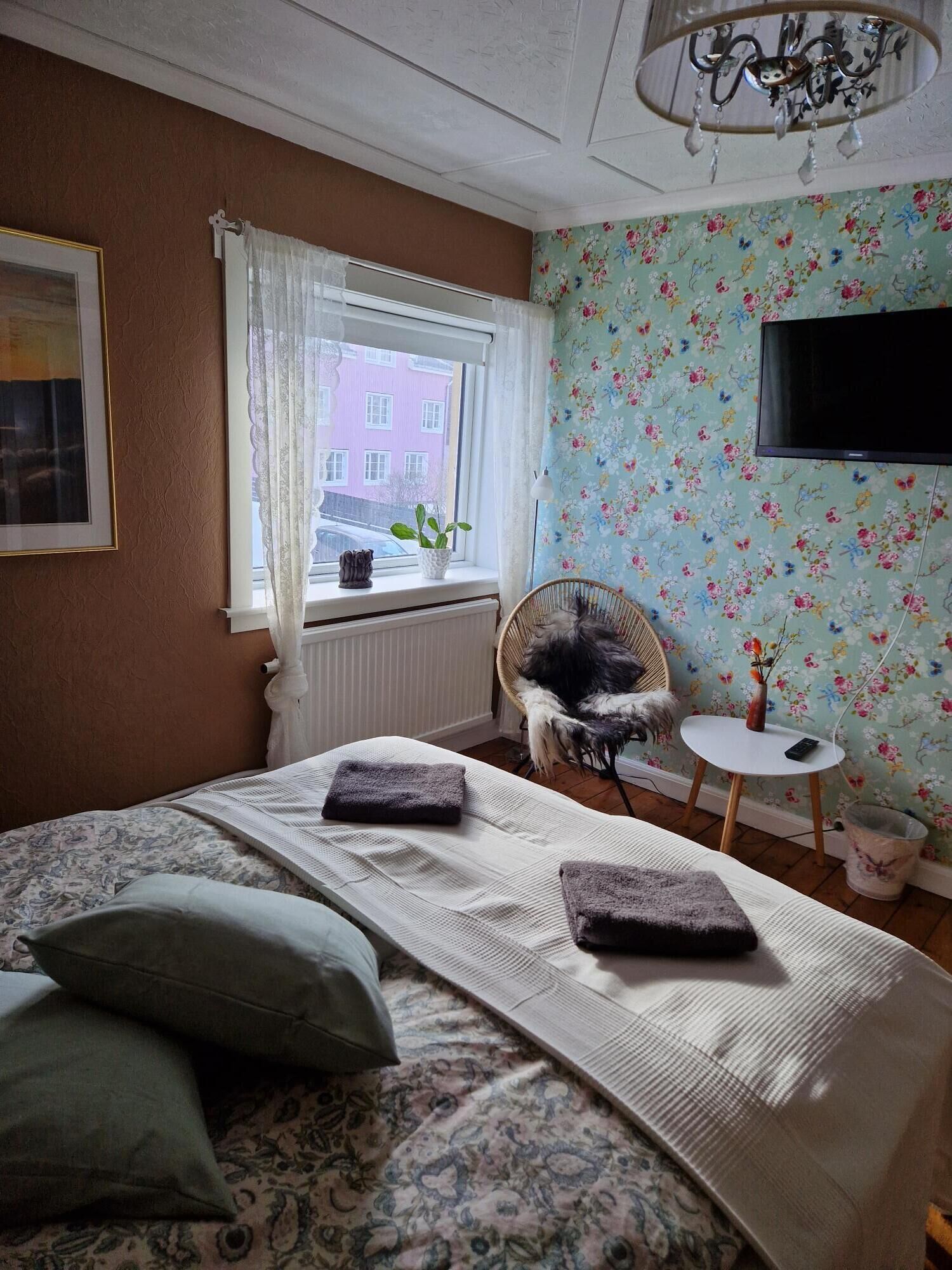A bedroom with a bed, a chair, a small table, a potted plant, and a window with a view of buildings.